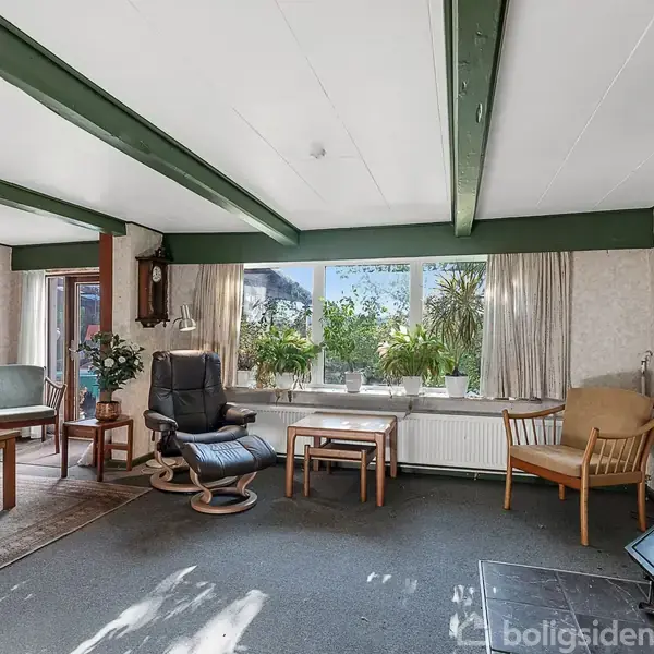 Stue med brændeovn i hjørnet, et stort vindue med planter, stole og sofabord i midten. Rummet er lyst med grønne loftsbjælker og en tæppebelagt gulvflade.