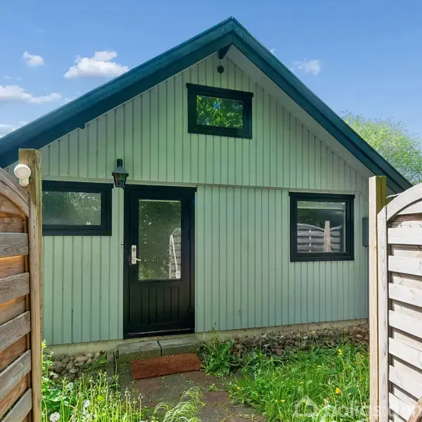 Et lille hus med en spids tagform, lysegrønt træpanel og sorte vinduer. Huset ses bag en havelåge af træ, omgivet af græs og buske under en blå himmel.