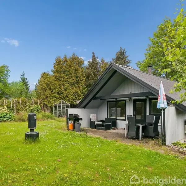 Et sort-hvidt sommerhus står i en grøn have med terrasse møbler og parasol. Omgivet af buske og træer under en blå himmel.