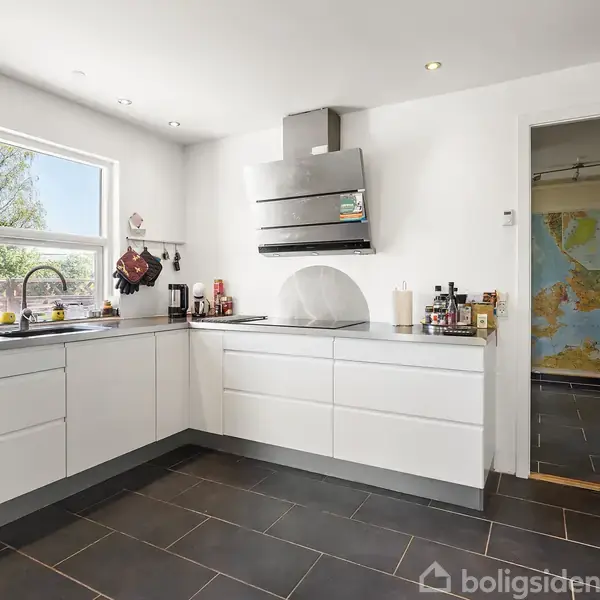 Køkken med hvide skabe under en stålbordplade, hvor der er køkkenredskaber og krydderier. Et vindue til venstre slipper lys ind. Slidstærkt mørkt flisegulv.