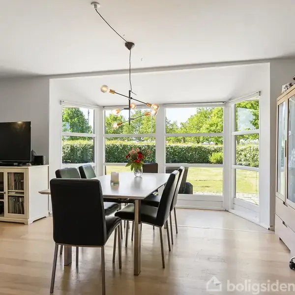 Spisebord med sorte stole står centralt, dekoreret med blomster. Store vinduer viser en grøn have. Et TV står til venstre, og et skab ses til højre.