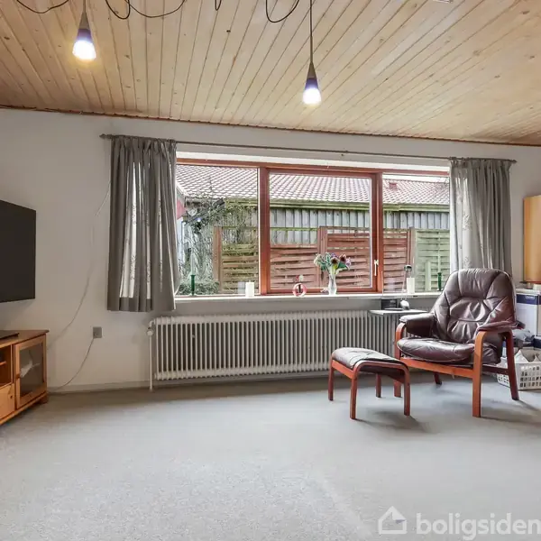 En stue med et mørkt læderlænestol og fodskammel står foran et stort vindue med gardiner. Et fjernsyn er placeret på et møbel til venstre.