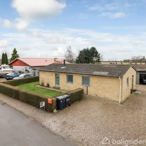 Et enplans hus står stille ved en grusbelagt indkørsel. Baggrunden inkluderer en rød bygning og landligt landskab. En hæk omkranser en lille forhave. Skraldespande står ved vejen.