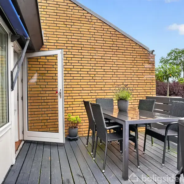 Terrassemøbler står på et moderne opbygget trægulv. Et sort bord med seks stole er placeret foran en murstensvæg. Åben glasdør tillader indgang fra huset, omgivet af grønne træer.