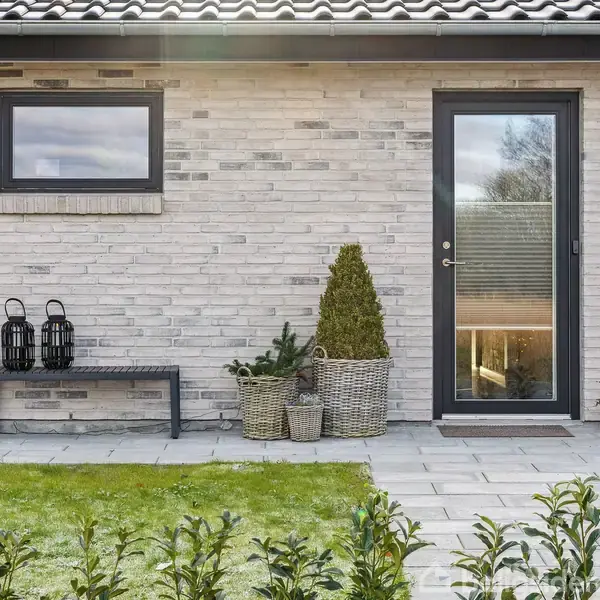 En muret husfacade med grå dør og vindue. Ved siden af står en bænk med sorte lanterner. Planter i kurve er placeret ved siden af døren. En græsplæne i forgrunden.