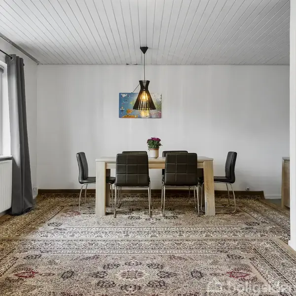 Spisebord med fire sorte stole; en vase med blomster på bordet. Rummet har hvidt loft, lyse vægge, tæppe på gulvet og et vindue med grå gardiner.