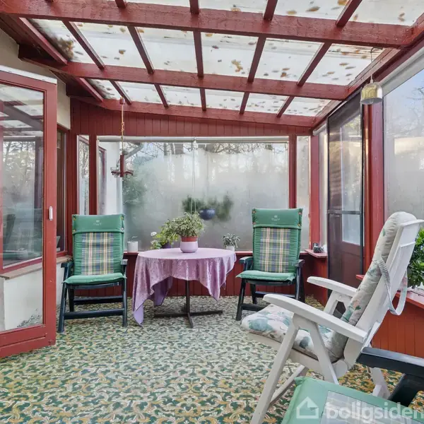 En glasterrasse har flere grønne og hvide stole omkring et rundt bord dækket med en lilla dug. Udsigten er delvis skjult af dug på glasset, mens mange planter tilføjer en naturlig stemning.