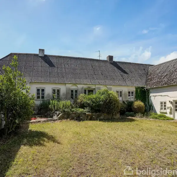 Gammel gård med hvidkalkede vægge og skråt, sort tag. I forgrunden ses en åben gårdsplads omgivet af bygninger, planter og buske med blå himmel i baggrunden.