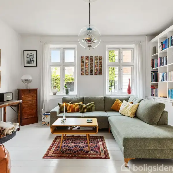 Grøn sofa står i et lyst stuerum med træhylder fyldt med bøger. Kaffebord foran sofaen og kunst på væggene skaber en indbydende atmosfære. Store vinduer giver naturligt lys.