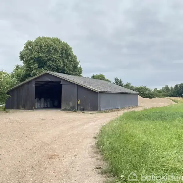 Et stort gråt lagerhus står åbent og tomt på en grusvej, omgivet af grønne marker og træer under en overskyet himmel. Der ses nogle sække inde i bygningen.