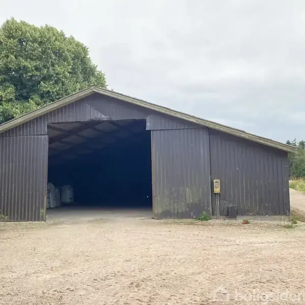 En stor, åben lade står på en grusvej omgivet af grønne træer og marker.