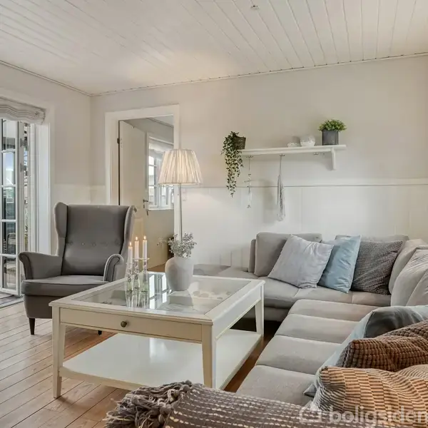 Stue med stor hjørnesofa fyldt med puder ved et sofabord. En lænestol står ved glasdøren til en terrasse. Hylder med planter pynter en væg, og der er naturligt lys.