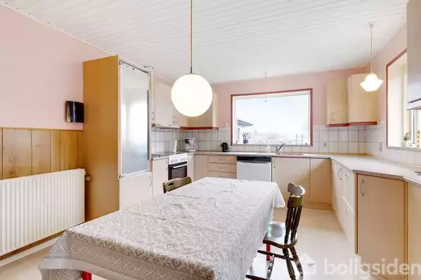 Et køkken med et bord dækket af en dug. Stole står omkring bordet. Der er lyse skabe, en opvaskemaskine, ovn og et stort vindue, der giver udsigt.