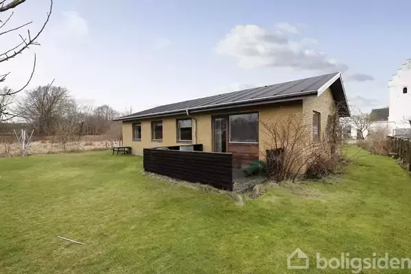 En enkeltstående murstenshus med solpaneler på taget står i en velholdt have. Huset har store vinduer og en overdækket terrasse. Haven er græsbevokset med nogle buske omkring.