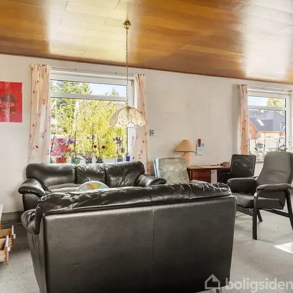 En stue med sorte lædermøbler, inklusiv en sofa og stol, placeret foran to store vinduer. Til højre er der et skrivebord med en lampe.