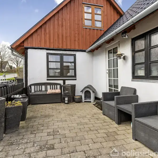 En hyggelig terrasse med kurvemøbler og plantekrukker. Møblerne står på flisebelægning foran et hvidt hus med en træoveretage. Der er også en lille hundeseng ved døren.