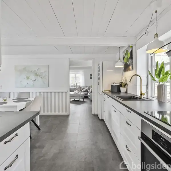 Køkken med hvide skabe og sort bordplade, indeholder kogeplade og vask. På bordet står en plante og køkkenmaskine. I baggrunden ses en spisestue med sofa og maleri.