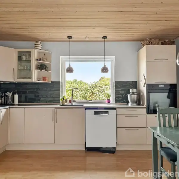 Køkken med hvide skabe og en opvaskemaskine midt for. Et stort vindue med udsigt til træer bagved. Til højre står et spisebord og stole.