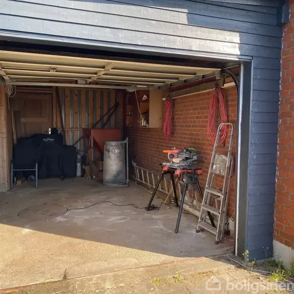 En garage med åben dør indeholder en stige og værktøjer, herunder en sav på et stativ. Væggene er af mursten med ophængte røde kabler til højre.