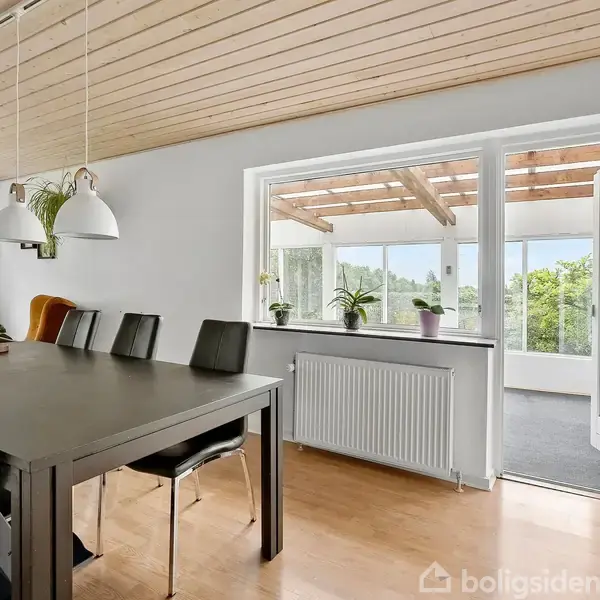 Veranda med store vinduer og mørkt tæppe, der giver udsigt til en have med træer og buske. En hvid bil ses parkeret udenfor, i nærheden af en grusvej.