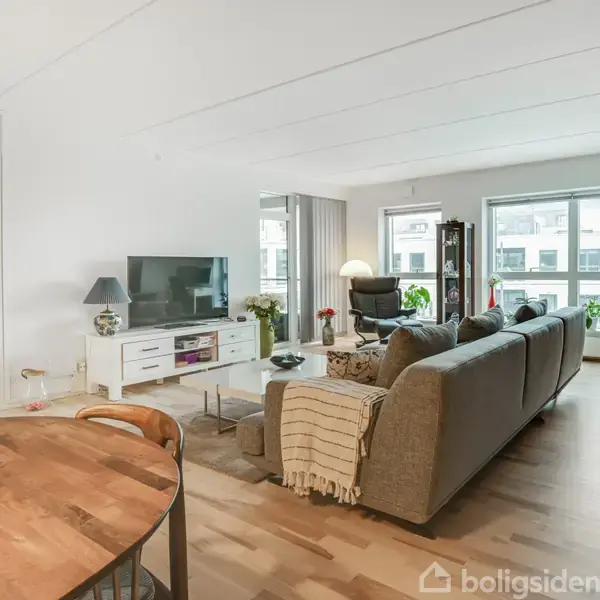 En stue med en grå sofa i midten, foran et tv på et hvidt mediemøbel. Store vinduer bagved lader lys ind. Rundt spisebord i forgrunden, omgivet af stole.