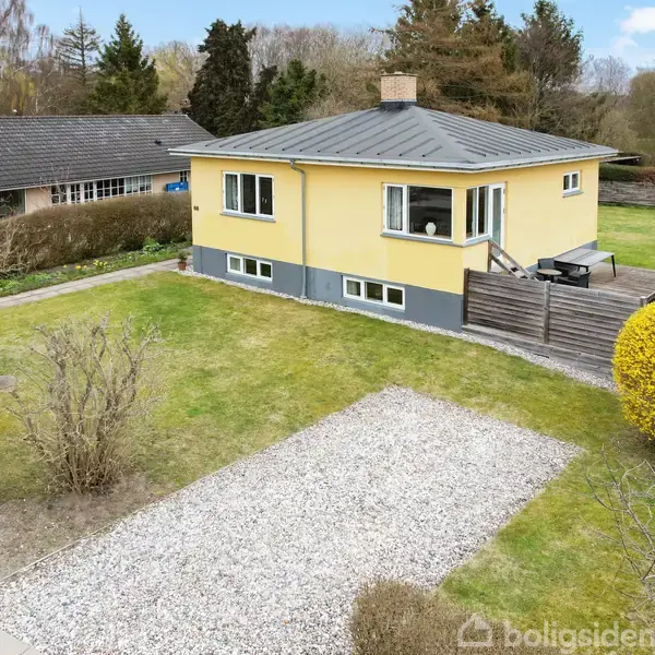 Et gult hus står på en grøn græsplæne med en grusbelagt indkørsel. Huset har en træterrasse med havemøbler og er omgivet af buske og træer i et villakvarter.
