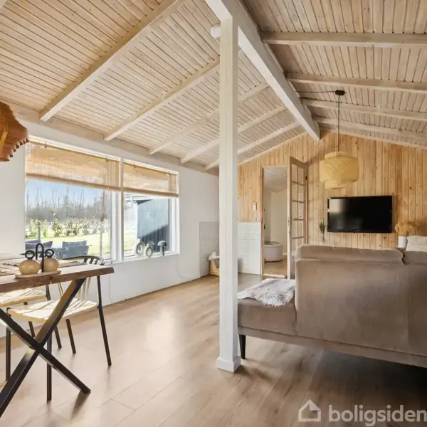 Træbord med stole står opstillet i en lys stue med træloft og gulv. Sofaen og væghængt TV ses i baggrunden. Store vinduer viser en have udenfor.