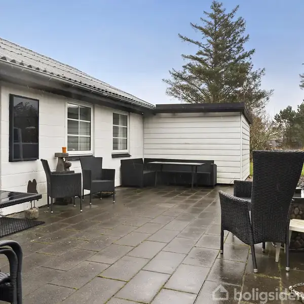 Terrasse med flisebelægning, hvor flere sorte kurvemøbler og et lille bord er placeret. Til højre ses et plankeværk, og der er høje træer i baggrunden.