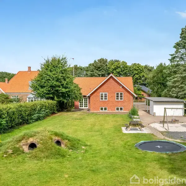 Rød murstenshus med hvidrammede vinduer står i baggrunden. Foran huset er en grøn have med en indbygget trampolin, legeplads og to græsklædte jordhuler. Baggrunden har træer og blå himmel.