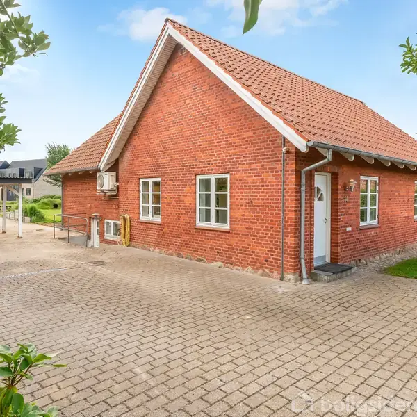 Rødt murstenshus står stille på en brolagt gårdsplads omgivet af grønne træer. Huset har et skråt tag og flere vinduer. Der er en carport til venstre for huset.