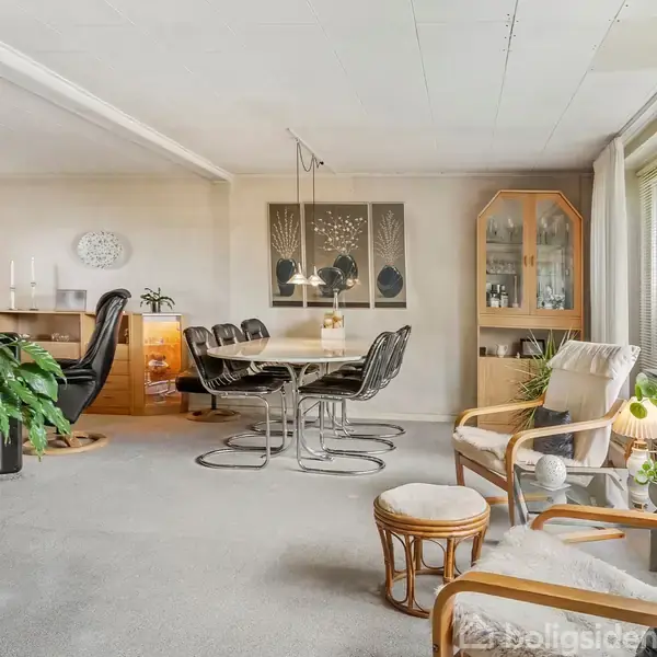 En lys stue med et rundt spisebord omgivet af stole, placeret nær store vinduer. Der er også en lænestol, planter og en reol i rummet, som er indrettet med lyse nuancer.