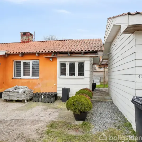 En ensidig orange murstensbygning med hvide vinduer står til venstre. Til højre er en hvid træbygning. Foran er der grus og græs. Tekst: "10".