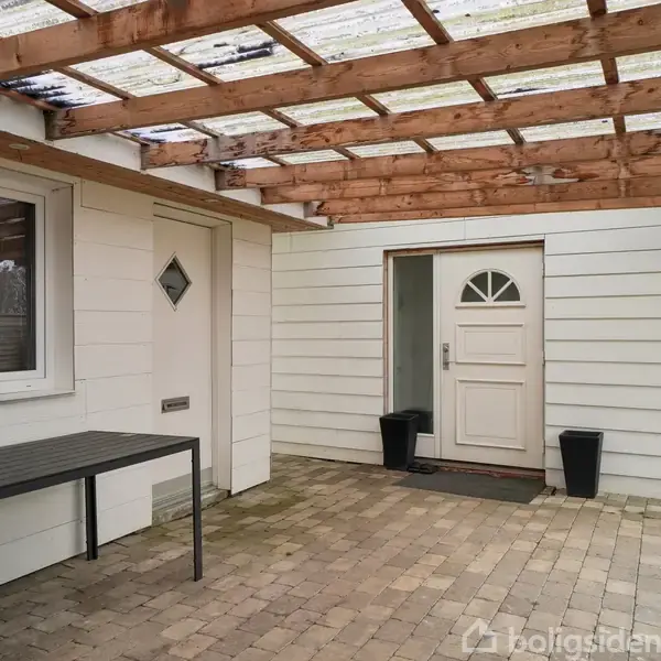 En overdækket terrasse med flisebelægning. Til venstre står et sort bord med stole, og til højre ses en hvid dør med bueformet vindue. Væggene er lysebrædder.