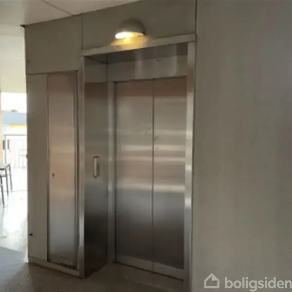 Elevator i rustfrit stål står stille i en opgang nær en balkon med stole og borde. Væggene er røde mursten, og en lampe lyser over elevatoren.