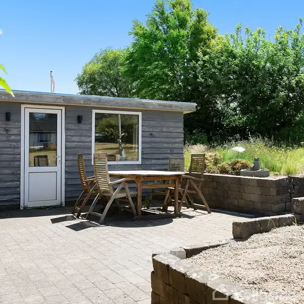 En havepavillon står på en flisebelagt terrasse med et bord og stole foran. Omkring pavillonen er græsplæne og grønne træer i baggrunden under en klar blå himmel.