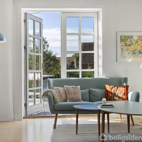 Sofa beliggende i en lys stue med åbne dobbeltdøre mod en altan. Der er et rundt sofabord, en gulvlampe og udsigt til grønne træer og huse udenfor.