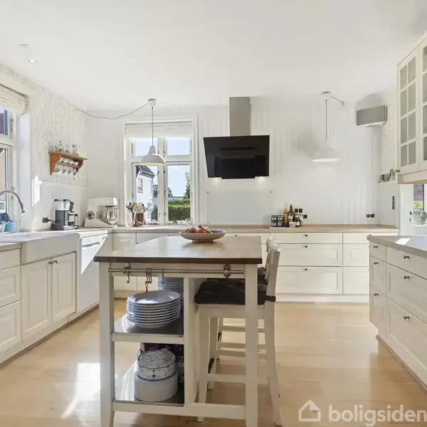 Et lyst køkken med hvidt interiør og træbordplader. En køkkenø står i midten med skåle og serveringsfade. Store vinduer lukker dagslys ind, og skabe tilbyder opbevaring.