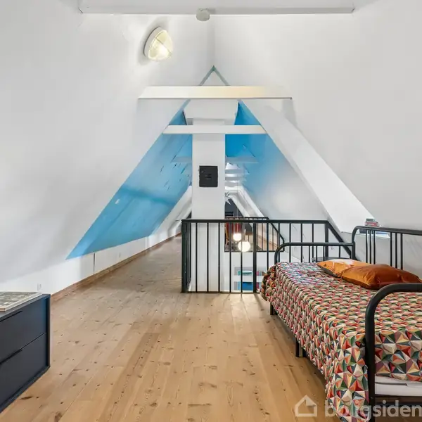 En hems med en enkeltseng med farverigt sengetæppe står ved en skrå væg. Trægulvet strækker sig mod en sort kommode med en lampe. Rummet er lyst og luftigt.
