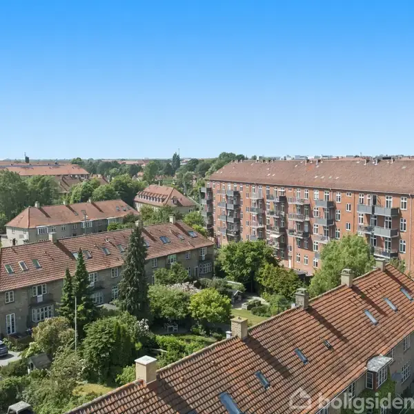 Udsigt over tagene af rækkehuse med røde tage i et boligområde. I forgrunden ses en altanræling, og i baggrunden strækker byen sig under en klar blå himmel.