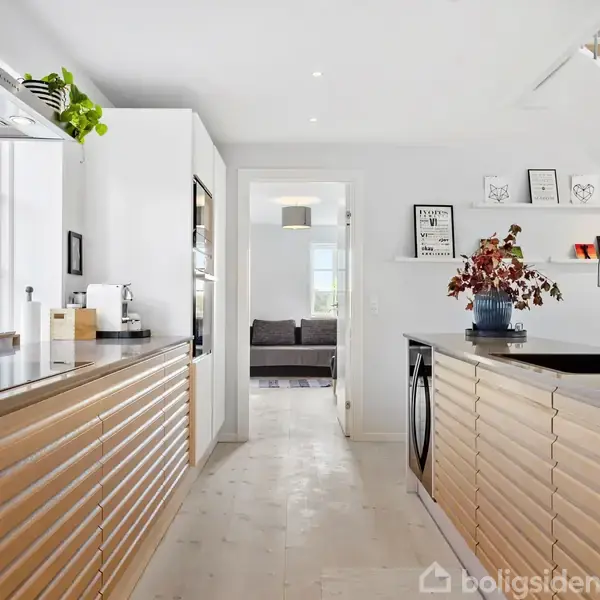 Et moderne køkken med træskuffer strækker sig langs begge sider. Planter og dekorationer pryder hylderne. I baggrunden ses en åbning til en stue med en sofa.