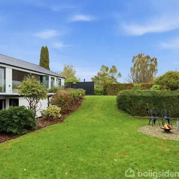 Et moderne hus står i en velplejet have. På græsplænen ses en rund brolagt terrasse med stole og en ildsted. Haven er omgivet af buske og træer under en klar blå himmel.