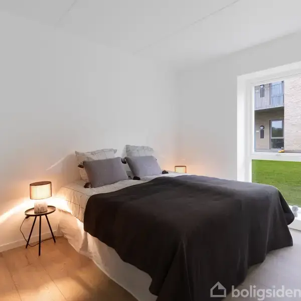 En seng med mørkt sengetæppe og puder står i et lyst soveværelse. Ved siden af er små natborde med lamper. Et vindue viser en græsplæne og et andet hus udenfor.