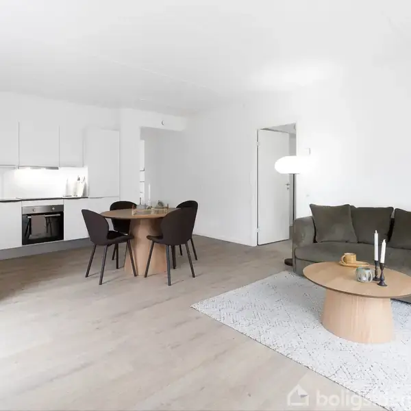 Grønt sofaarrangement står på et lyst gulvtæppe med lysestager og frugt på sofabordet. Til venstre er et indbygget hvidt køkken med spisebord og stole i en moderne stue.