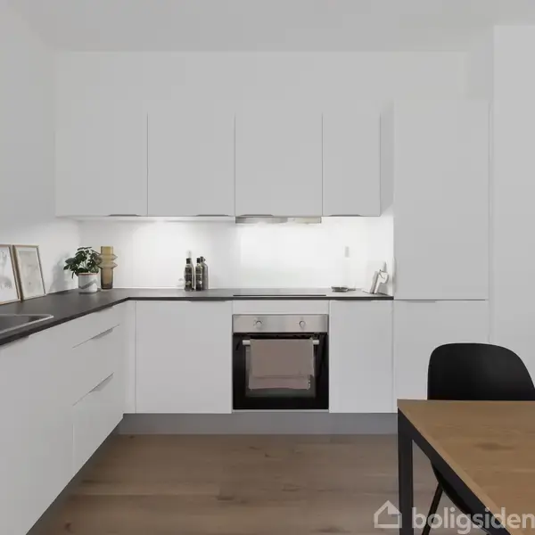Køkken med hvide skabe, en indbygget ovn, sort bordplade og vask. Væggen har dekorationer, og der er en træbordplade med en sort stol ved siden af.