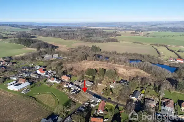En landsby set fra luften med en rød pil pegende mod et bestemt hus. Omgivet af marker, skovområder og spredte boliger i naturskønt terræn.