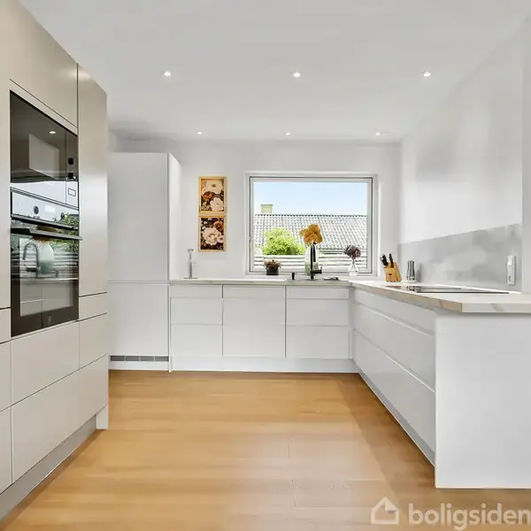 Et moderne køkken med hvide skabe og integrerede apparater. Rummet er lyst med et stort vindue. En plante står i hjørnet, og trægulvet strækker sig gennem rummet.