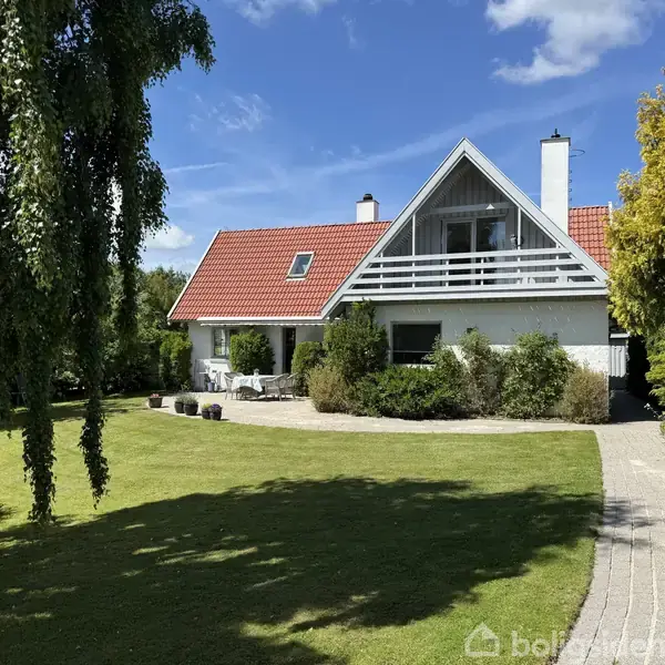 En vindeltrappe fører opad i en entré med hvide vægge og trægulv. Et træbord med blomster står til venstre, og en glasdør åbner ind til et lysere rum.