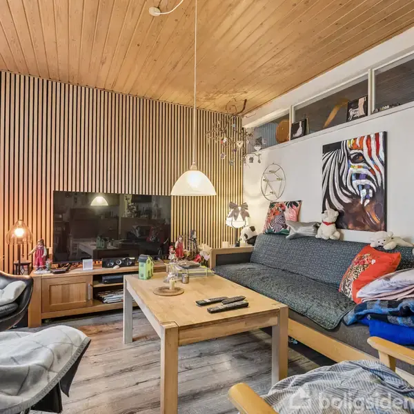 Stue med sofa fyldt med puder og tæpper, finde midt i et hyggeligt rum med træloft og stribede trævægge. Et TV står overfor, omgivet af lamper og dekorationer. En hængende lampe lyser sofabordet op.