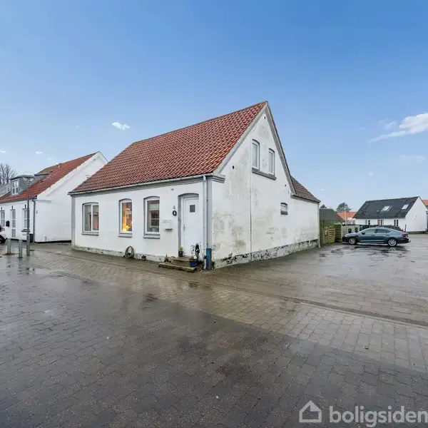 Hvidt hus med rødt tag står på brolagt plads. Bilen er parkeret til venstre. Området er åbent med andre huse i baggrunden under en blå himmel.