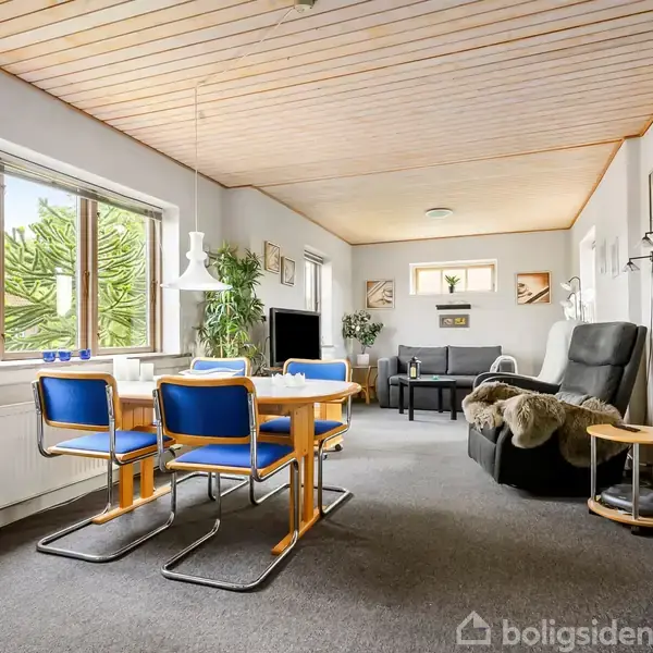 Spisestue med blå stole ved træbord, hvor sollys strømmer ind fra store vinduer. Opholdsstue i baggrunden med sofa og lænestol. Lyst træloft og malerier på væggene.